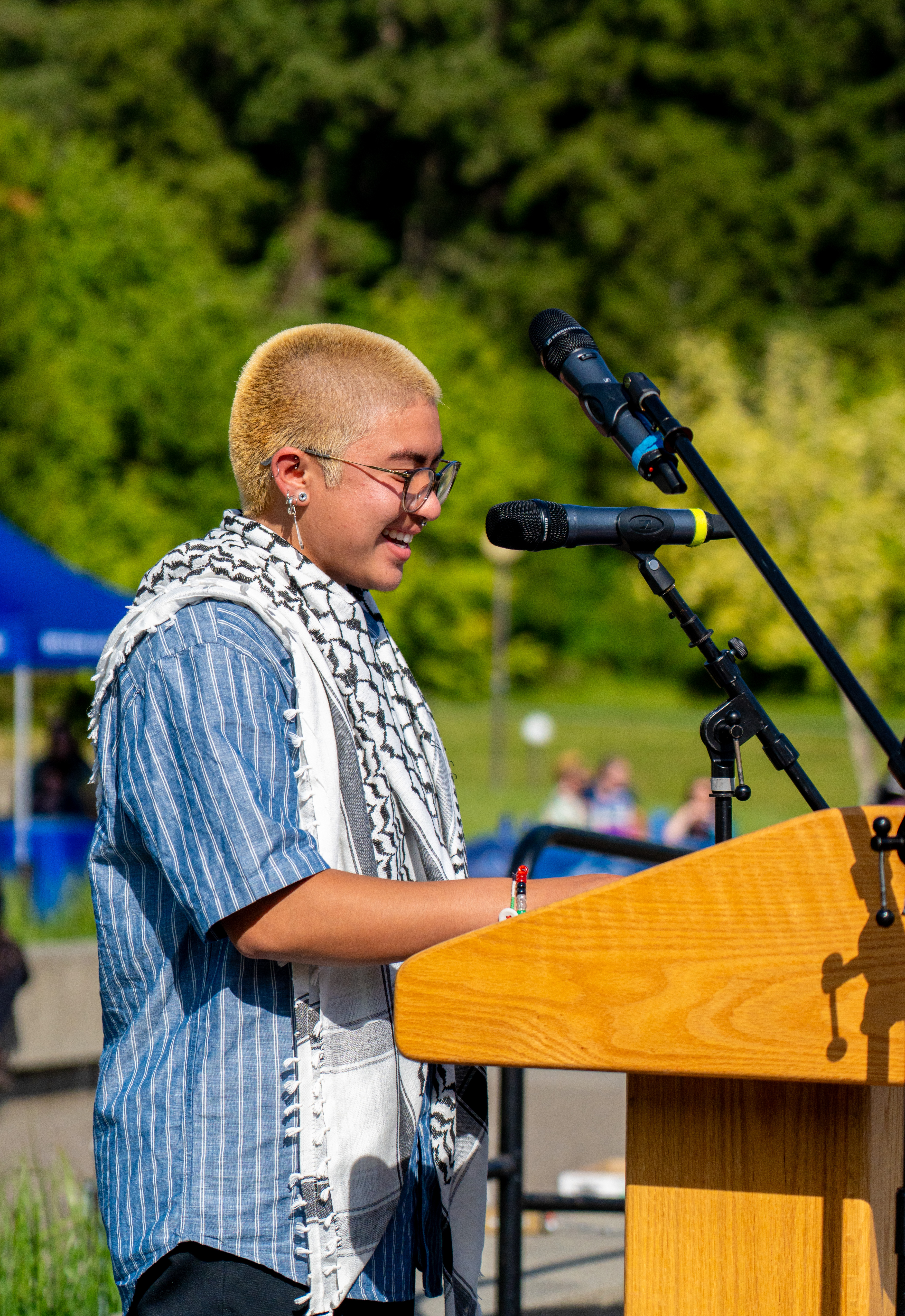 Sof Trujillo wearing a blue shirt and whte and black keffiyeh standing at the podium smiling.